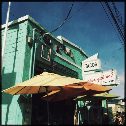 Colorful taqueria with a turquoise exterior and yellow umbrellas, featuring a sign that reads "TACOS" and "¿Por qué no?".