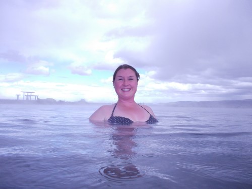 A smiling woman in a polka dot bikini stands in a misty hot spring, with a cloudy sky and distant structures in the background.