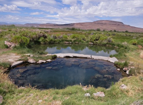 A serene natural hot spring surrounded by lush greenery and rocky terrain under a cloudy sky.