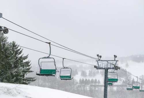 Ski lift chairs suspended in a snowy landscape, surrounded by trees and a foggy atmosphere.
