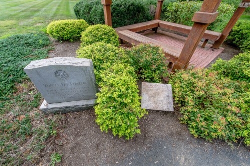 A stone memorial and plaque surrounded by neatly trimmed bushes near a wooden gazebo in a grassy area.
