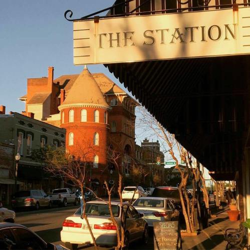 A street view featuring a sign for "The Station," with historic buildings and trees in warm evening light.