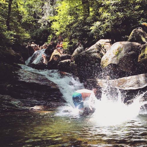 A person splashes in a clear stream surrounded by lush greenery and rocky terrain. Sunlight filters through the trees.