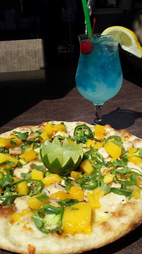 A colorful pizza topped with mango, chicken, and herbs, alongside a blue cocktail with a lemon slice and cherry.