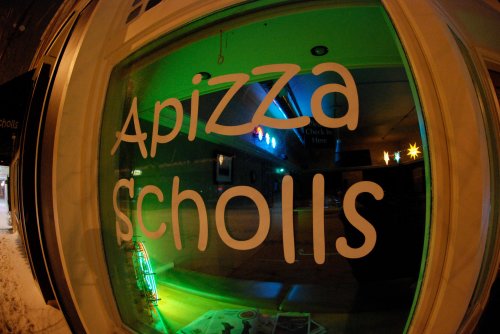 A brightly lit window displaying the name "Apizza Scholls" with colorful lights in the background.