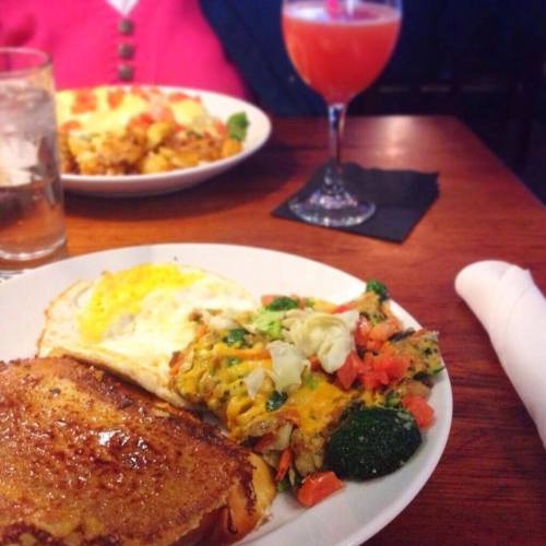 A plate with French toast, a fried egg, and a vegetable medley, alongside a glass of pink drink on a wooden table.