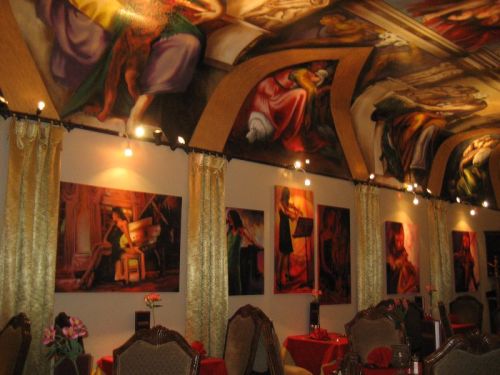 Interior of a restaurant featuring ornate ceiling murals and framed artwork on the walls, with elegant seating.