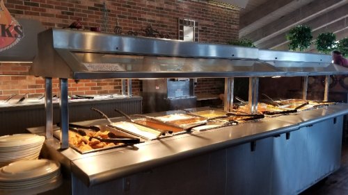 A buffet station with various food dishes under metal covers, set against a brick wall in a casual dining setting.