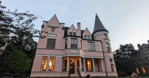 A large pink Victorian house with a turret, surrounded by trees, at dusk with warm light glowing from the windows.