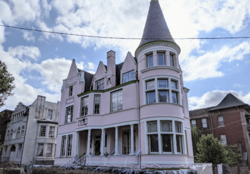 A large, pink Victorian-style house with a turret, surrounded by trees and a cloudy sky.