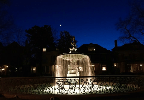 A illuminated fountain at night, surrounded by trees and historic buildings under a dark blue sky.