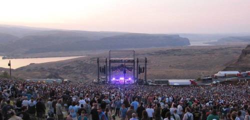 A large crowd at an outdoor concert, with a scenic landscape and river in the background during sunset.