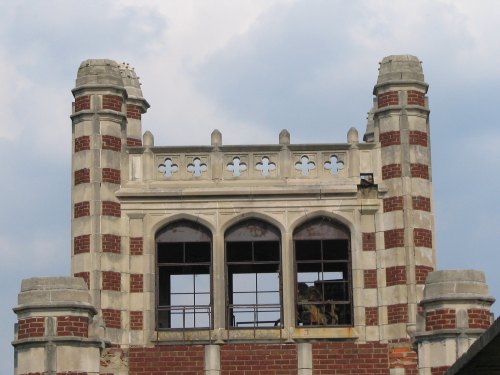 Historic building with red brick and stone accents, featuring arched windows and decorative towers against a cloudy sky.
