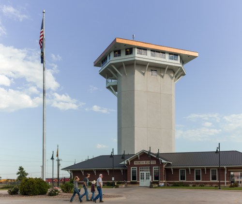 A tall observation tower beside a building, with two people walking in front and an American flag nearby.