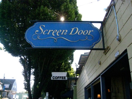 Sign for "Screen Door" in blue with decorative lettering, located near a coffee shop, surrounded by trees.