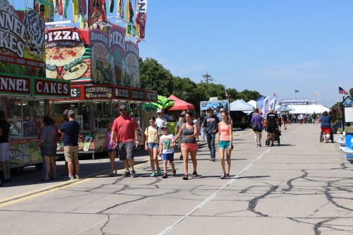 A lively fair scene with food stalls and people walking along a paved path under a clear blue sky.