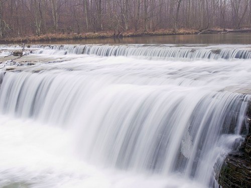 A serene waterfall cascading over rocks, surrounded by bare trees and a misty atmosphere.