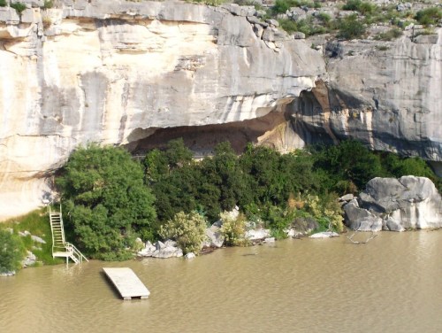 A rocky cliff with a cave entrance, surrounded by greenery, next to a calm river and a wooden dock.