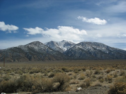 Snow-capped mountains rise against a blue sky with scattered clouds, surrounded by a dry, grassy landscape.