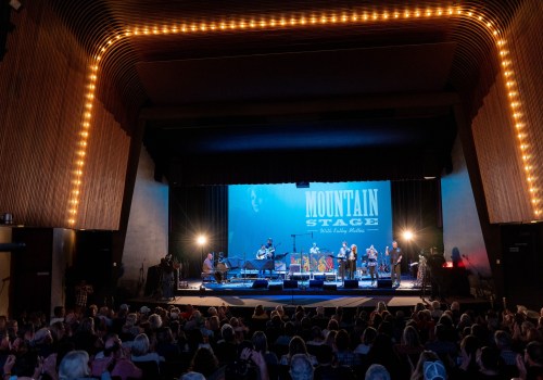 A live music performance on stage with a band, audience in view, and "Mountain Stage" backdrop illuminated.