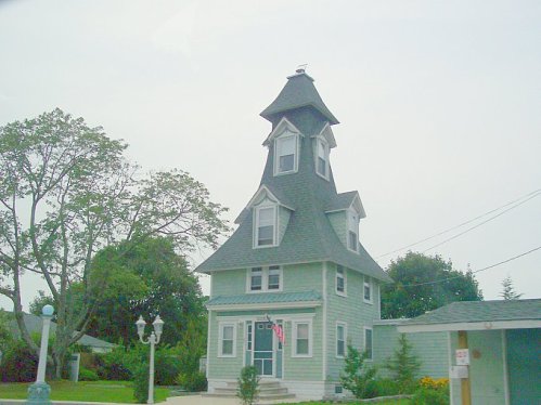 A unique green house with a tall, pointed roof and decorative features, surrounded by trees and a clear sky.