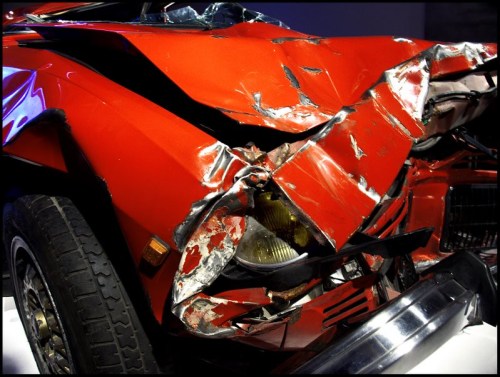A close-up of a severely damaged red car, showing crumpled metal and broken headlights.