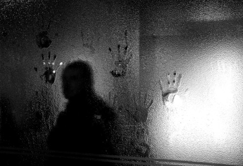 A shadowy figure stands behind a fogged glass, with handprints visible on the surface.