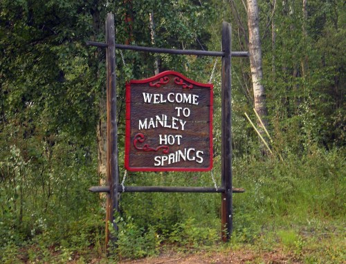 A wooden sign reading "Welcome to Manley Hot Springs" surrounded by greenery.