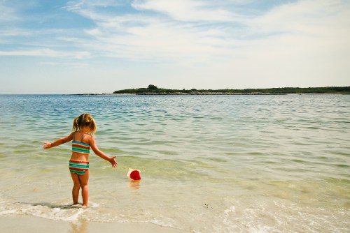 A young girl in a striped swimsuit plays at the beach, reaching for a red bucket in the shallow water.