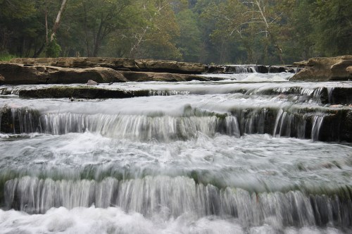 A serene waterfall cascades over rocky ledges, surrounded by lush greenery and trees.