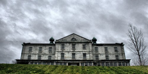 A large, abandoned building with a gray facade under a cloudy sky, set on a grassy hill.