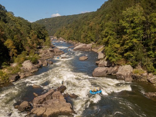 Aerial view of a river with rapids, surrounded by lush green trees and rocky banks, featuring a blue raft with people.