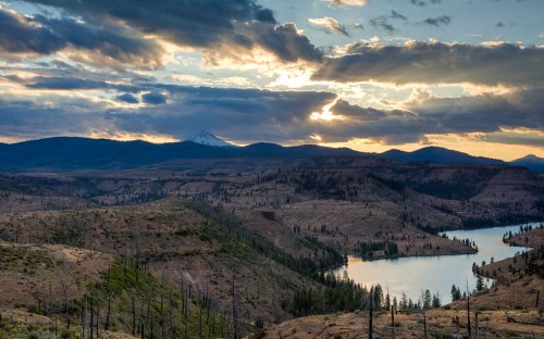 A scenic landscape featuring rolling hills, a river, and mountains under a cloudy sky at sunset.