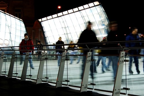 A bustling urban scene at night, with blurred figures walking past a modern glass structure.