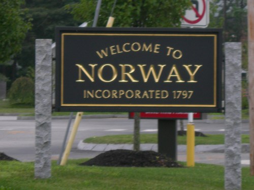 Sign welcoming visitors to Norway, established in 1797, with decorative stone posts and greenery in the background.