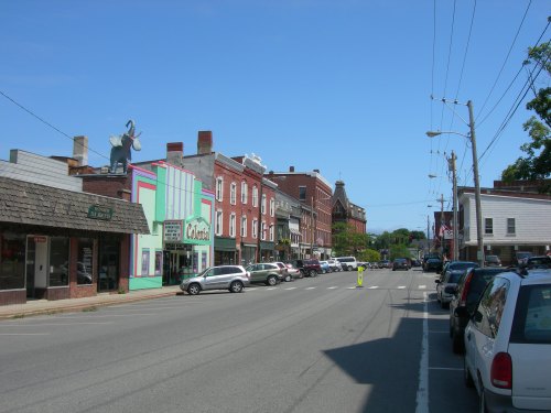 A small town street lined with shops and parked cars under a clear blue sky. A large elephant sign is visible on a building.
