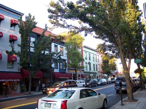 A street view featuring shops and trees, with parked cars and buildings in a quaint town setting.