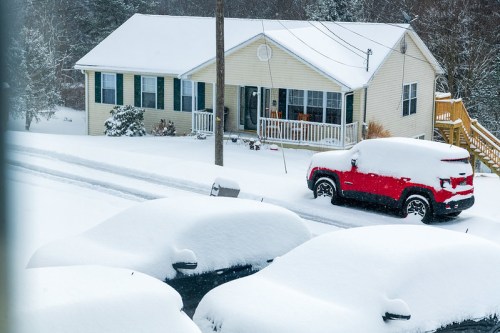 A snow-covered house with a red car and several buried vehicles in a winter landscape.