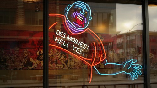 Neon sign of a cartoonish figure with the text "Des Moines: Hell Yes!" displayed in a window.