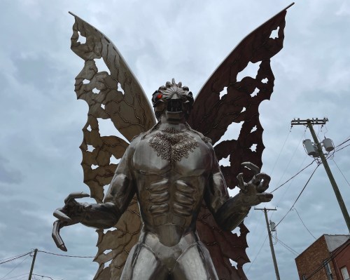 A large metallic statue of a creature with butterfly wings, standing against a cloudy sky.