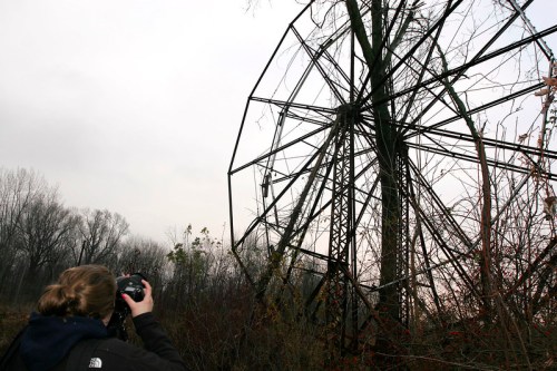 A person photographs a rusted Ferris wheel surrounded by overgrown vegetation on a cloudy day.