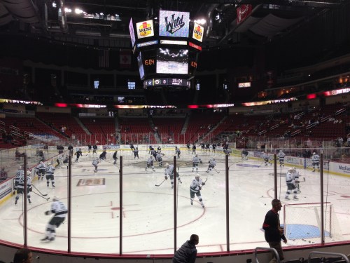 Hockey players warm up on the ice in a nearly empty arena, with a large scoreboard overhead.