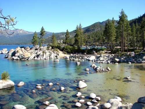 A serene lake surrounded by rocky shores and tall pine trees, with clear blue water reflecting the sky and mountains.