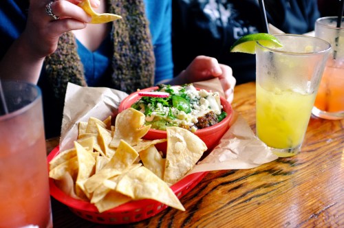 A person holds a chip above a basket of nachos topped with cheese and herbs, with drinks on the table.
