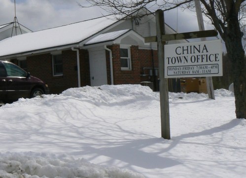 A snowy scene featuring a brick building with a sign for the China Town Office, indicating office hours.