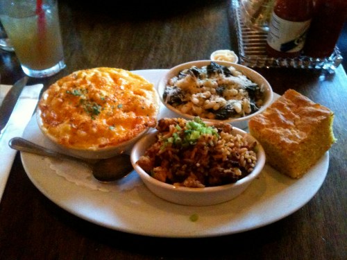 A plate with macaroni and cheese, greens, spicy rice, and cornbread, served with a spoon and a drink in the background.