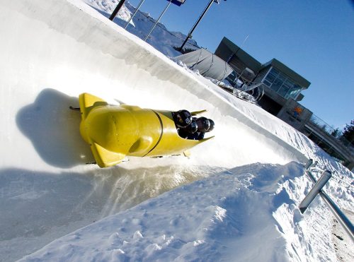 A yellow bobsled speeds down a snowy track, with two athletes inside, under a clear blue sky.