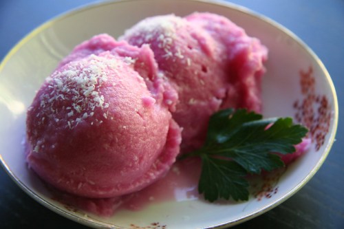 Two scoops of pink sorbet topped with shredded coconut, served in a decorative bowl with a sprig of parsley.