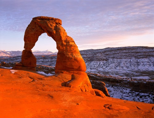 A natural stone arch stands against a colorful sky, surrounded by snow-covered terrain and rocky landscapes.