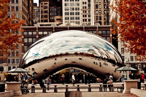 A reflective, bean-shaped sculpture in an urban park, surrounded by autumn trees and city buildings.